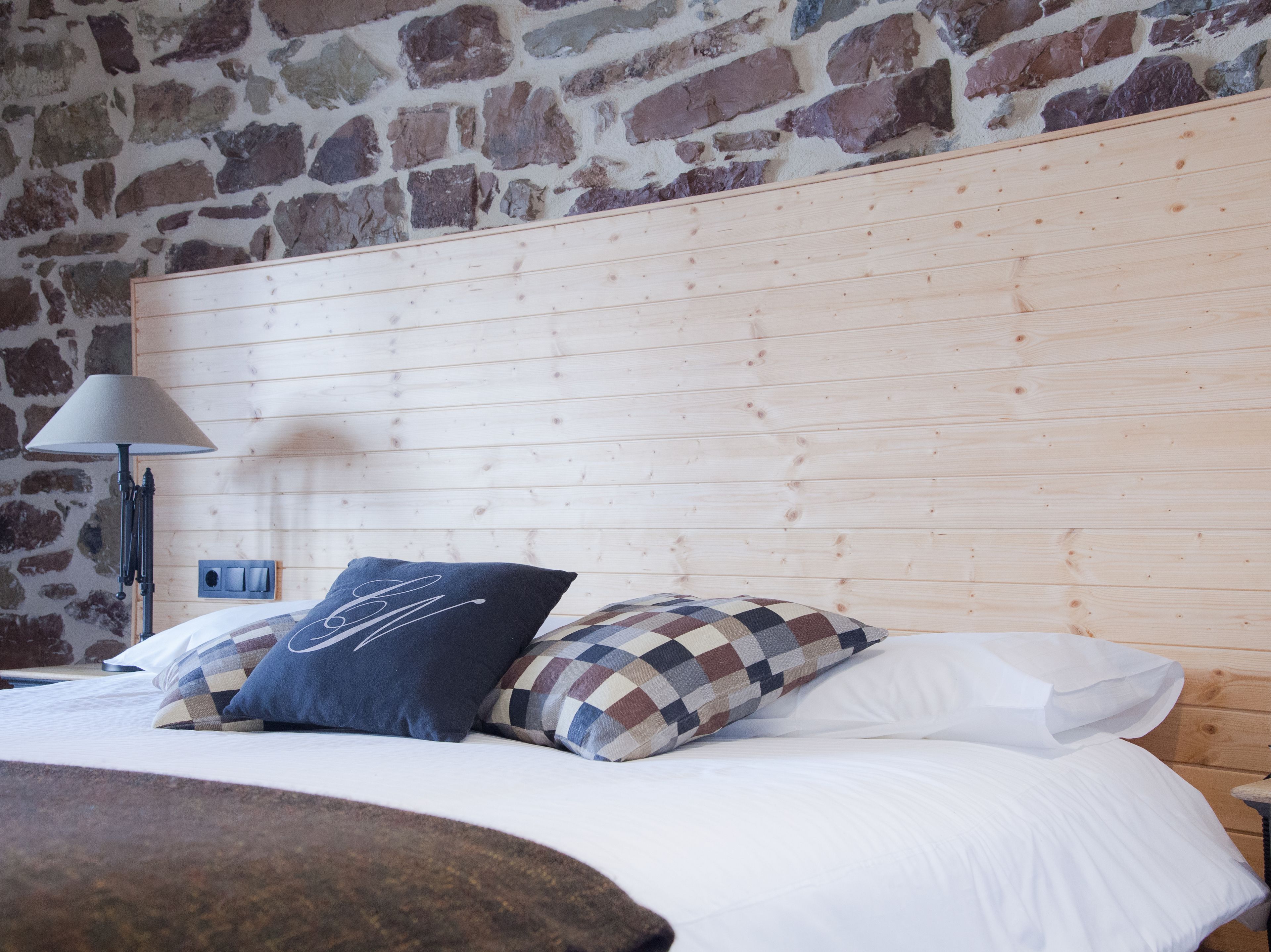 Un lit avec du linge de lit blanc, une tête de lit en bois et deux oreillers, dont l’un est à carreaux et l’autre porte un monogramme.