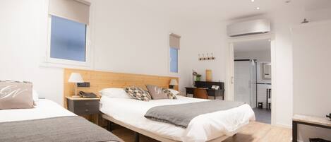 A modern hotel room with two beds, a wooden headboard, bedside tables with lamps, a small desk, and a view into the kitchen area.