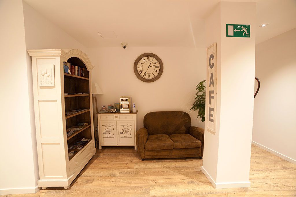 Una habitación con piso de madera, un sofá marrón, un armario blanco con libros, un reloj y un cartel que dice "CAFE".