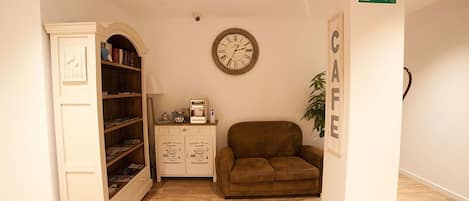 A room with a wooden floor, a brown sofa, a white cabinet with books, a clock, and a sign that says "CAFE".