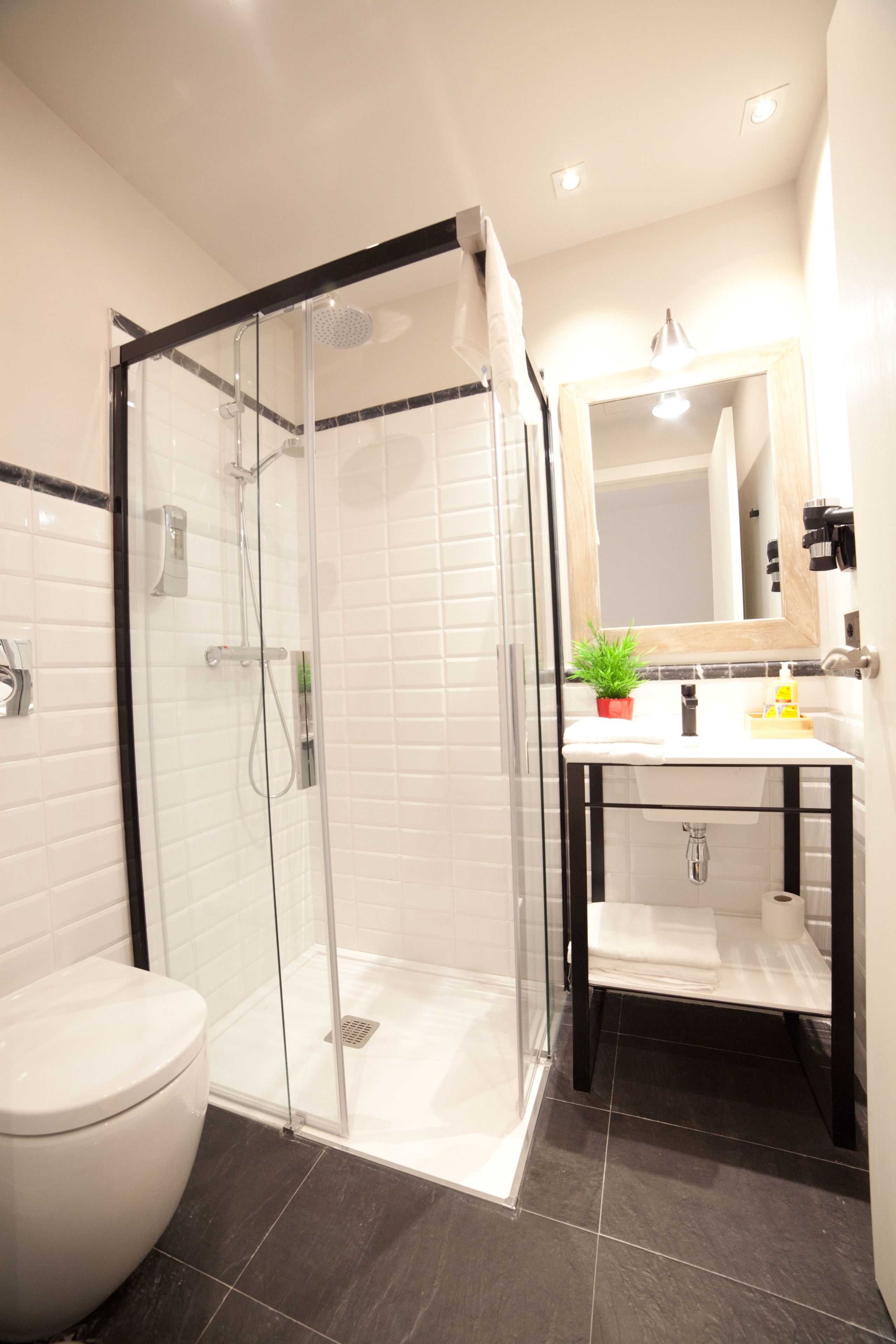 Un baño moderno con ducha envuelta en vidrio, inodoro blanco, lavamanos con espejo arriba y piso de baldosas oscuras.