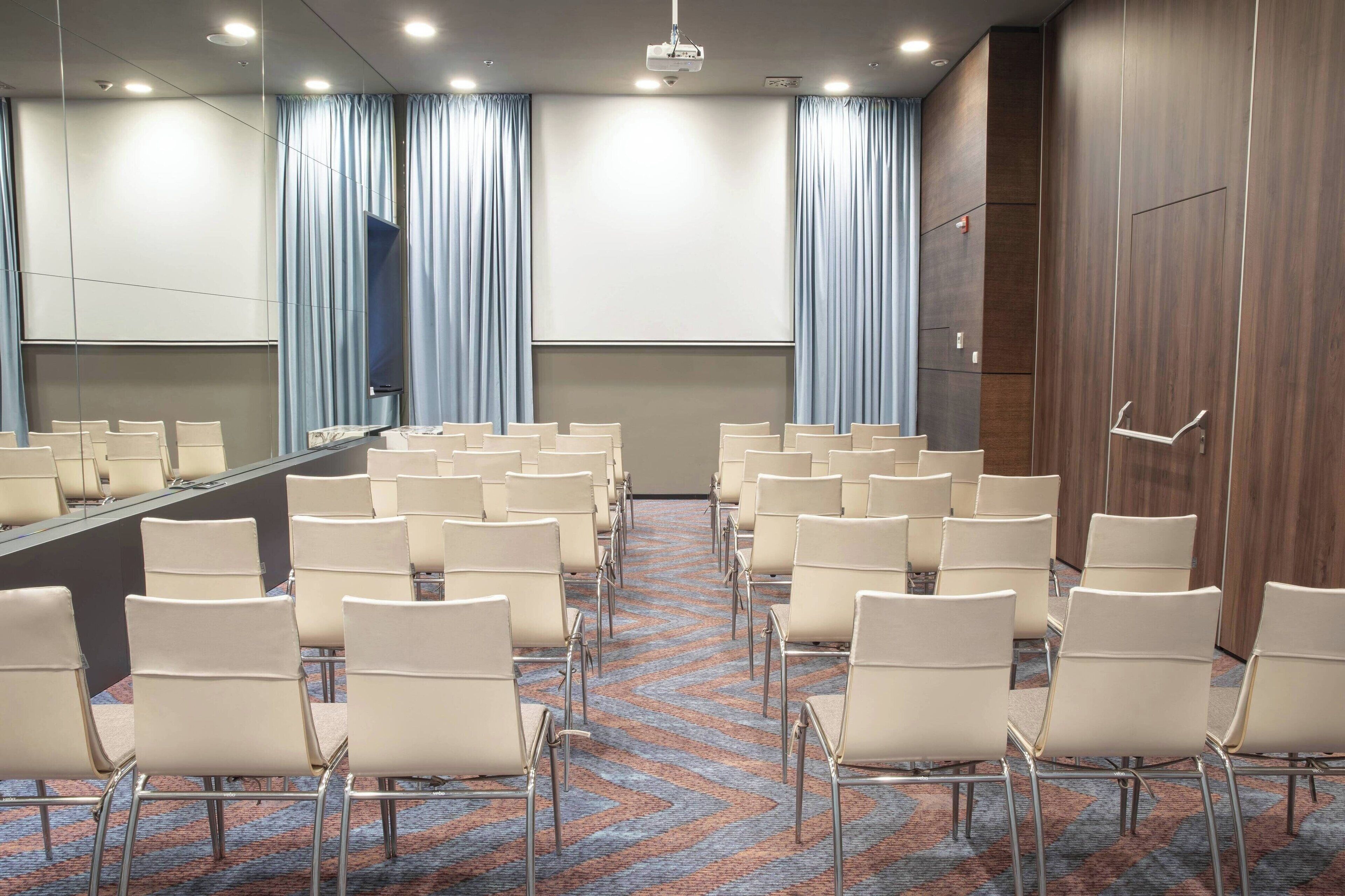 Ein Konferenzraum mit Reihen von Stühlen, die ein großes Tisch und einen Bildschirm gegenüberstehen, mit Vorhängen und einer Wand aus Holzverkleidung.