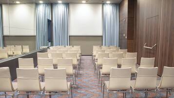 Ein Konferenzraum mit Reihen von Stühlen, die ein großes Tisch und einen Bildschirm gegenüberstehen, mit Vorhängen und einer Wand aus Holzverkleidung.