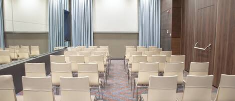 Un salón de conferencias con filas de sillas orientadas a una gran mesa y una pantalla, cortinas y un muro de paneles de madera.