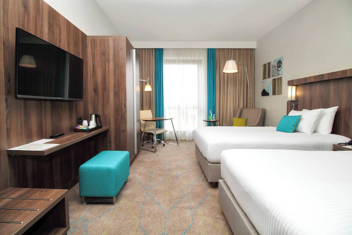 A hotel room with two beds, a desk, a chair, a television, and a window with curtains.