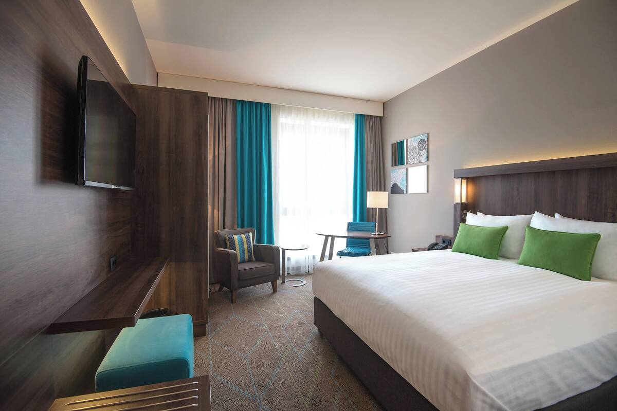 A hotel room with a large bed, a desk, a chair, a small table, and a TV mounted on the wall.
