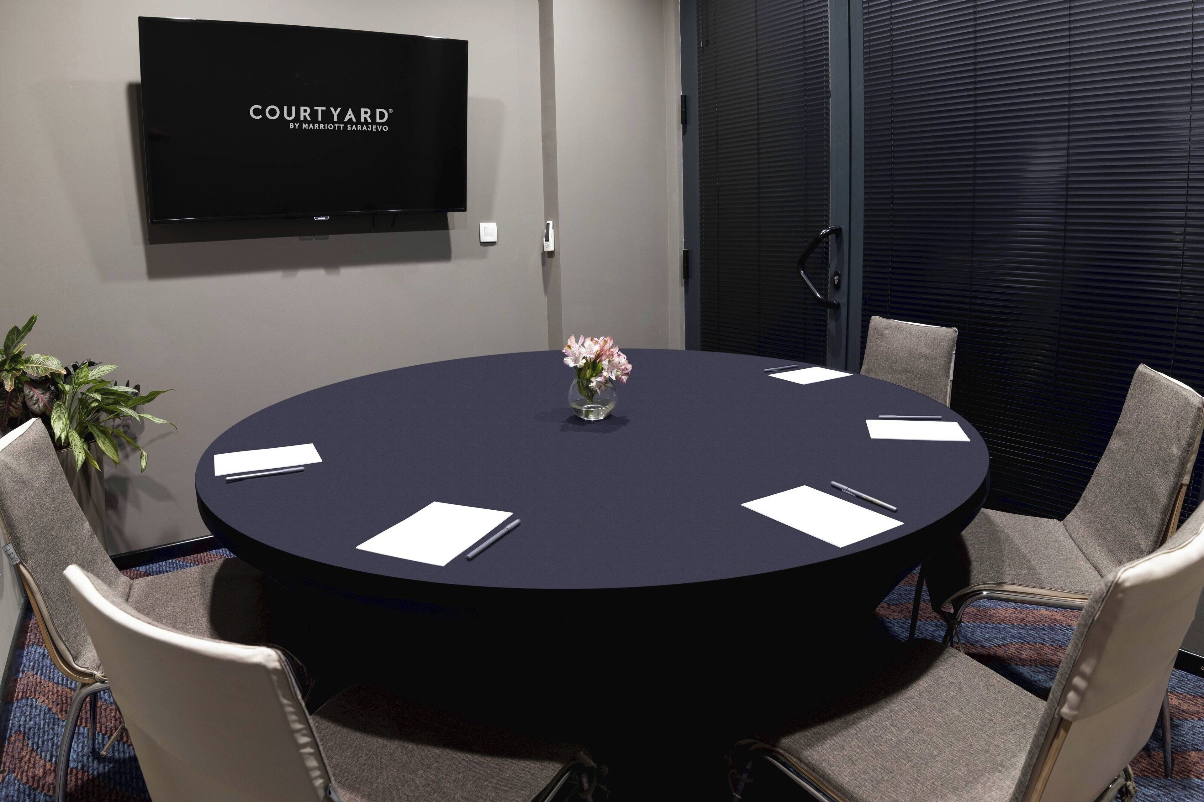 A conference room with a round table, six chairs, a TV on the wall, and a potted plant.