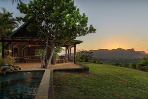 Un pabellón de madera con piscina, rodeado de exuberante vegetación y vistas a montañas distantes.