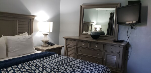 A hotel room with a bed, a nightstand with a lamp, a dresser, a mirror, and a TV mounted on the wall.