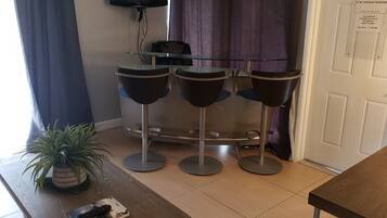 Una habitación con una mesa de vidrio, cuatro sillas negras, una planta en maceta, un control remoto y un televisor sobre un soporte.