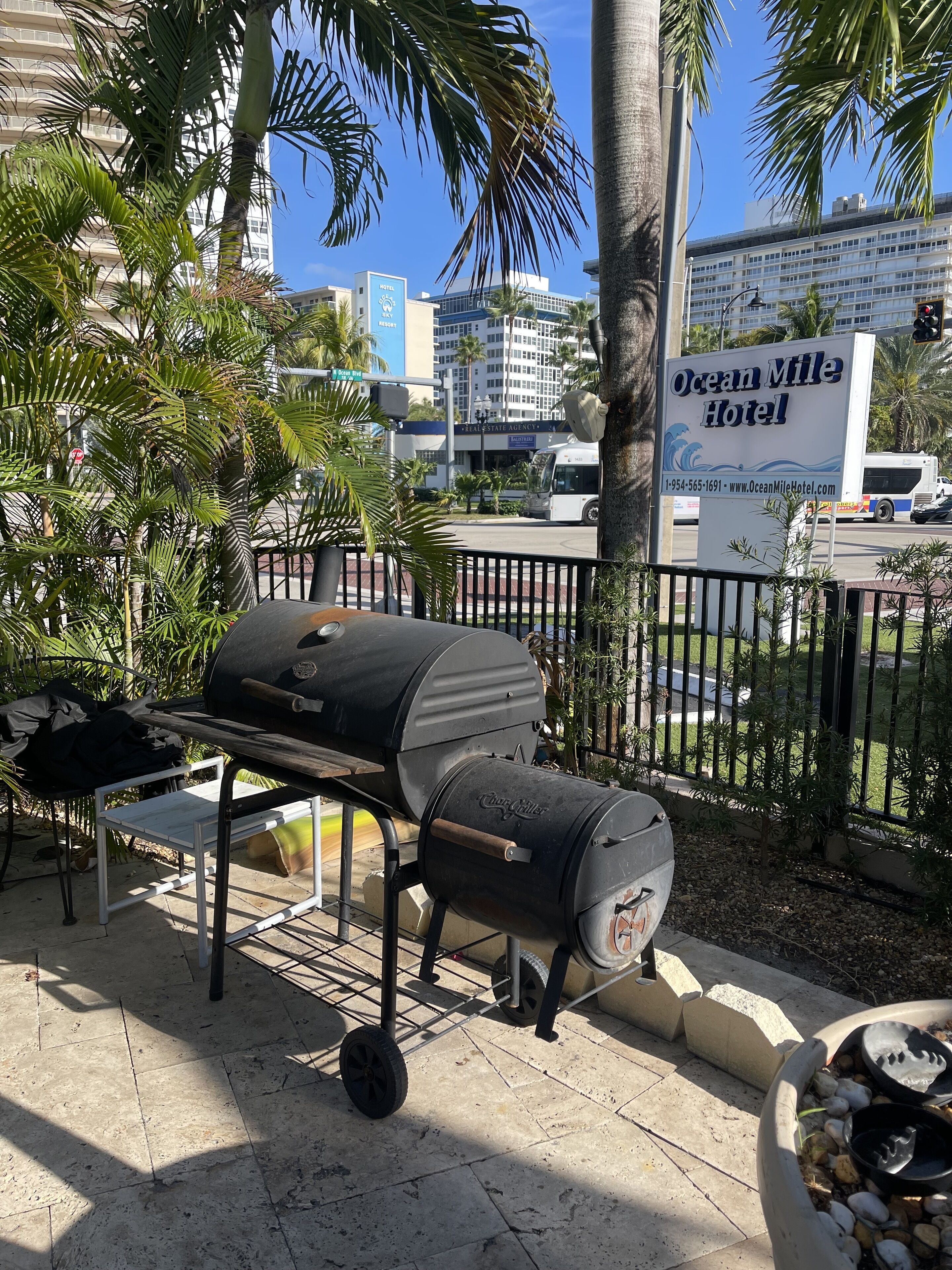 An outdoor grill area with a barbecue grill, a table, and a chair. In the background, there is a sign for the Ocean Mile Hotel and a bus.