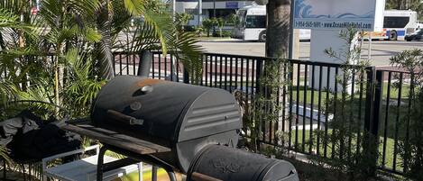 Uma área de churrasco ao ar livre com churrasqueira, uma mesa e uma cadeira. Ao fundo, um letreiro do Hotel Ocean Mile e um ônibus.