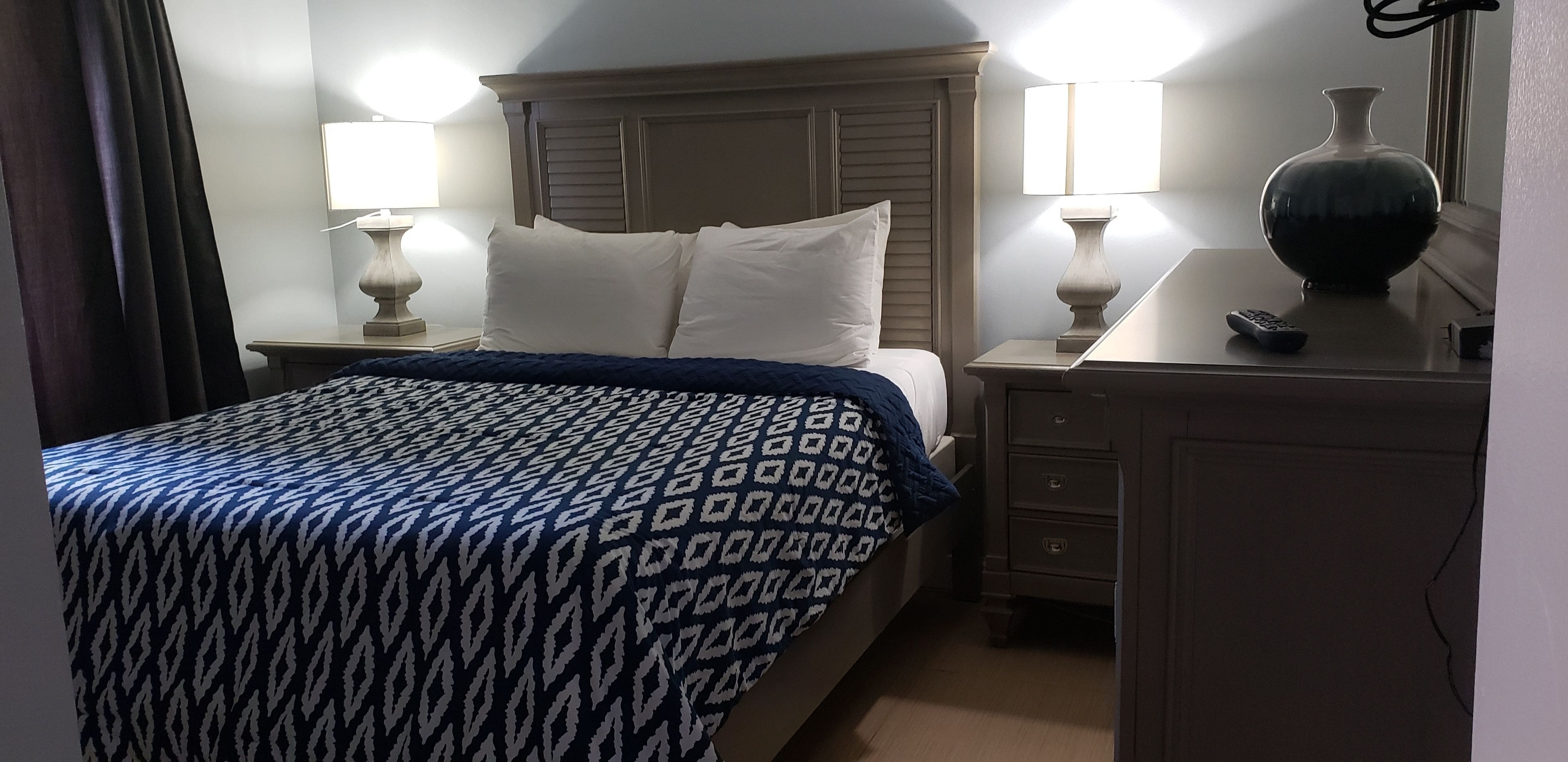 Una cama bien hecha con una colcha estampada, dos lámparas de noche, un jarrón en una mesita y un control remoto.