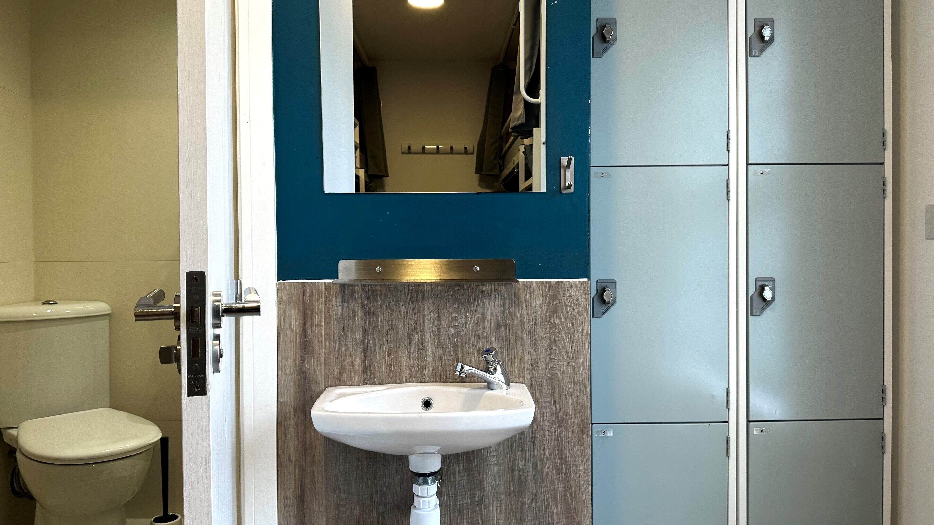 A bathroom with a toilet, sink, and a mirror above the sink. There are lockers on the right side.