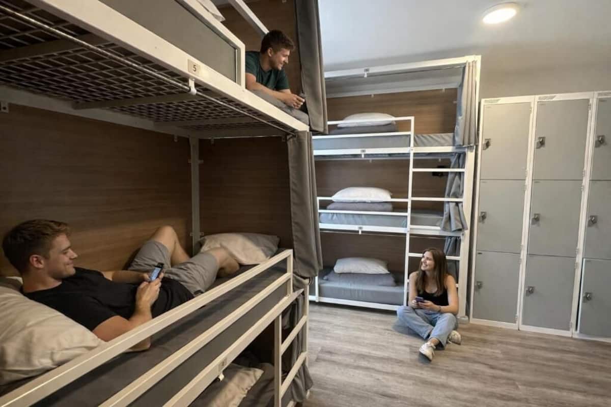 A bunk bed room with a person lying on the lower bunk, another person sitting on the floor, and a third person standing on the upper bunk.