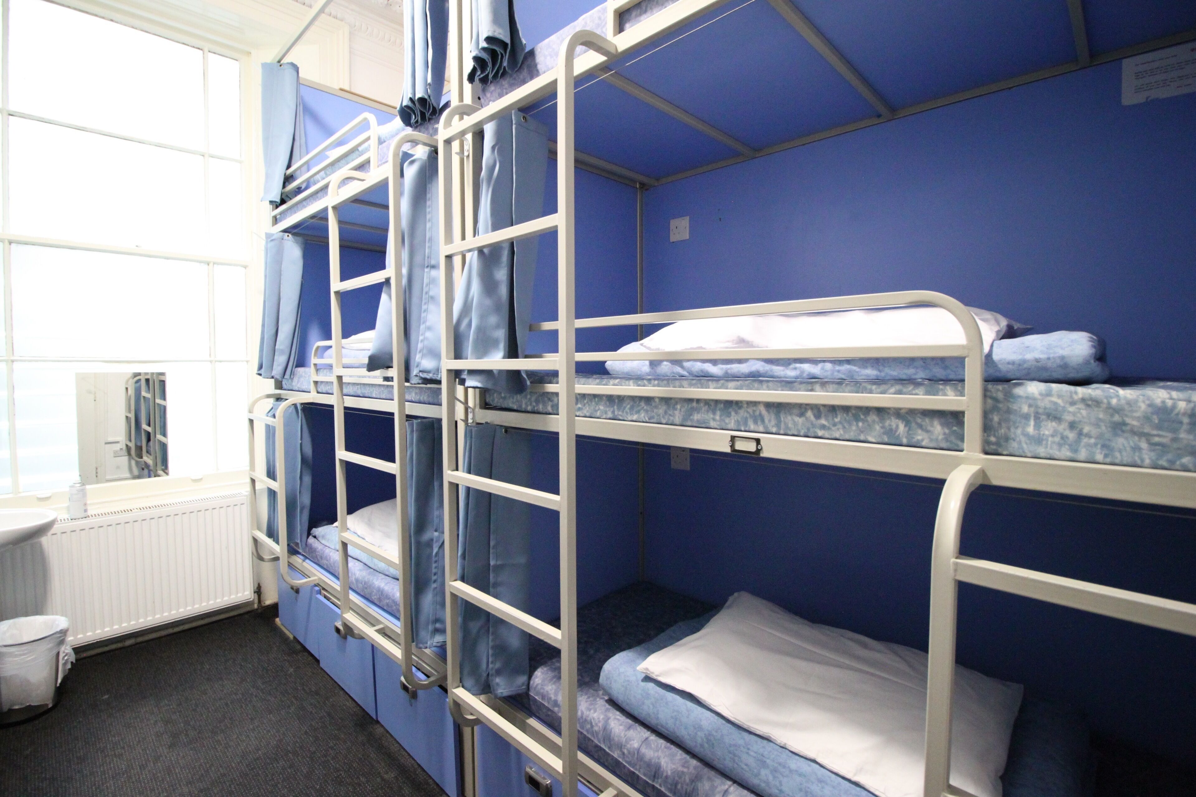 Une chambre avec un lit superposé, une fenêtre, un radiateur et une petite poubelle blanche.