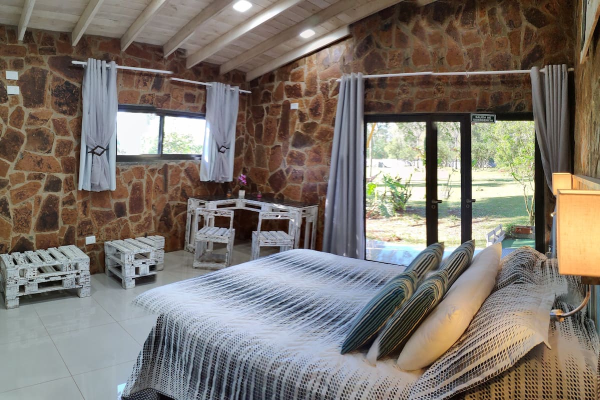 A rustic room with a stone wall, a bed with a white bedspread, two white chairs, and a table.