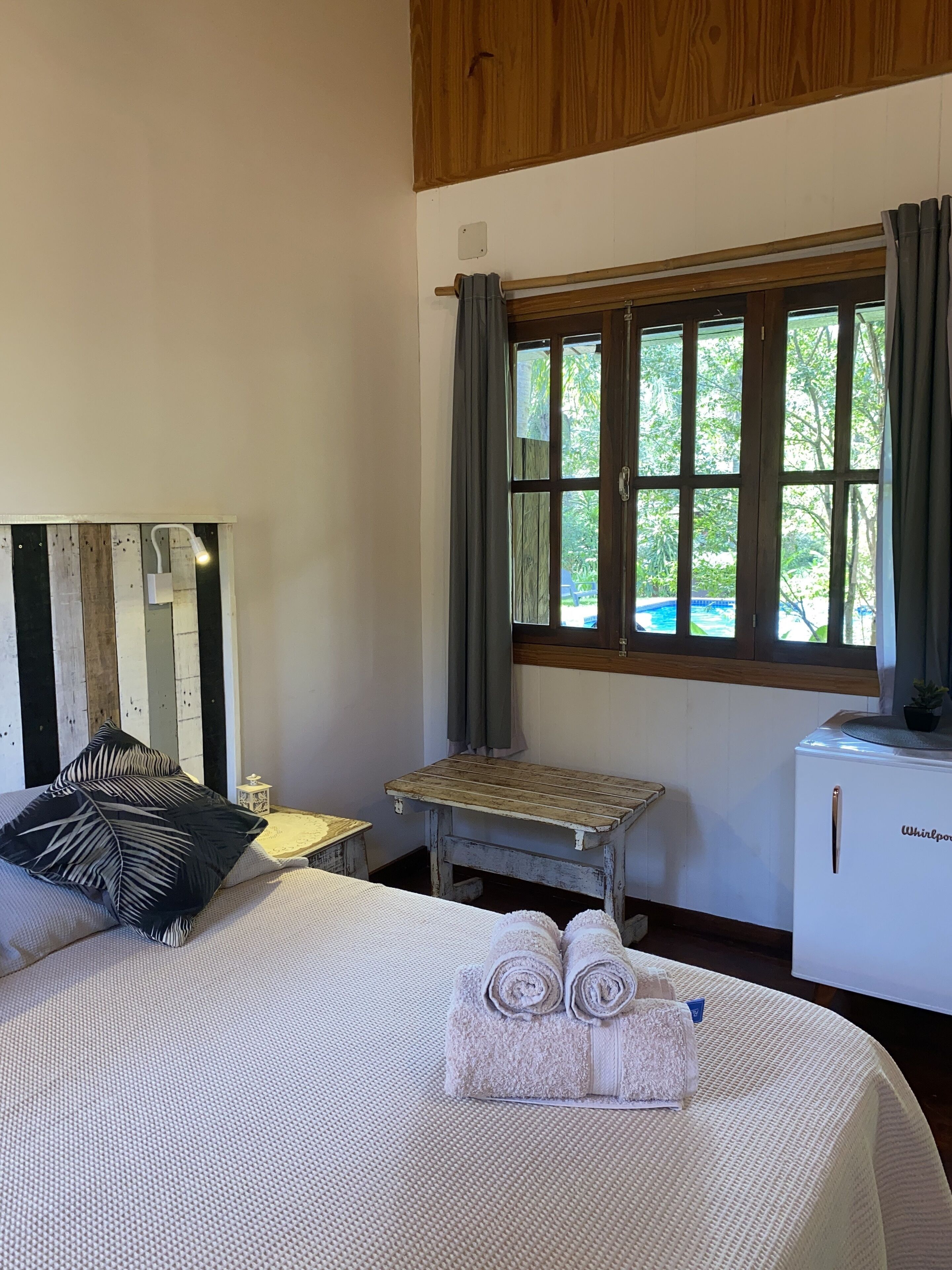 Un dormitorio con cama, una mesita, una ventana con cortinas y un refrigerador.