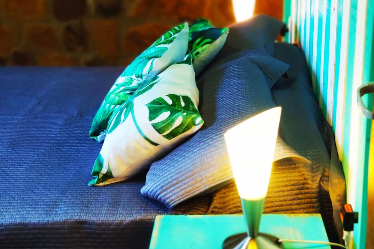 A bed with a green pillow and a blue blanket, a bedside table with a lamp, and a stone wall in the background.