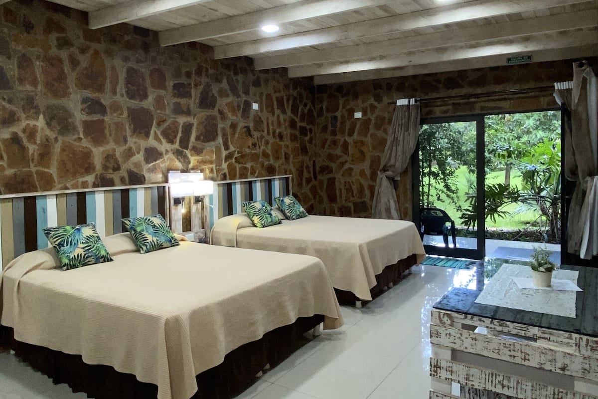 A room with two beds, stone walls, a tiled floor, and a view of greenery outside.