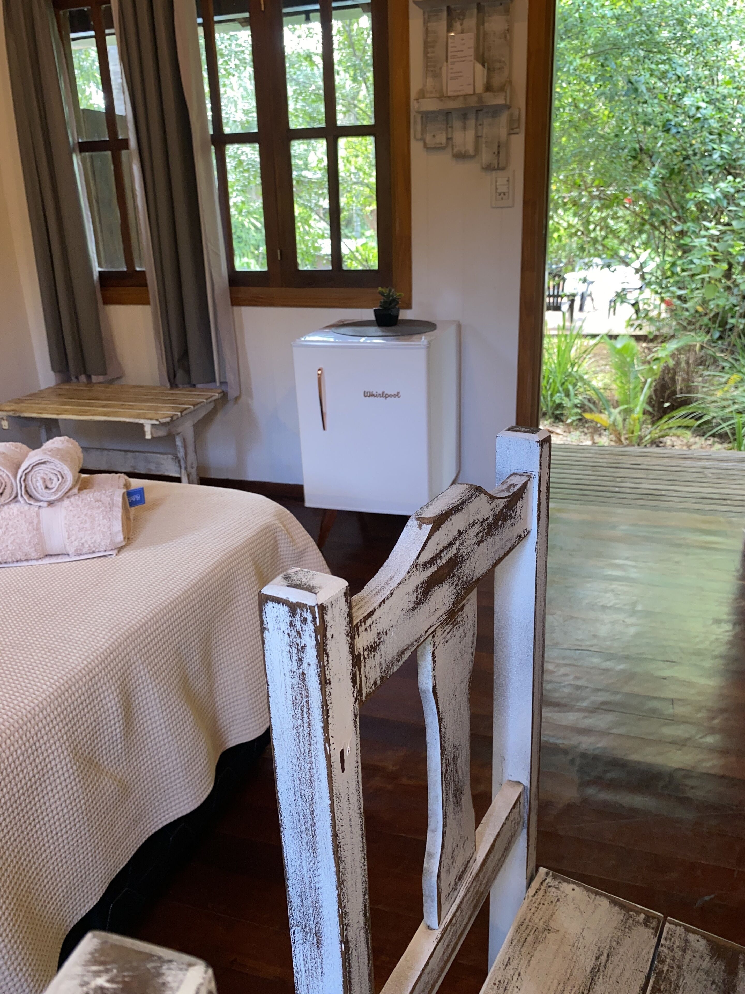 A rustic room with a bed, a small table with towels, a refrigerator, and a view of greenery outside.