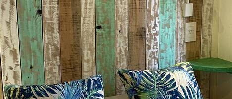A room with a rustic wooden headboard, two tropical-patterned pillows, a woven-texture sofa, and a small green shelf with a light fixture.
