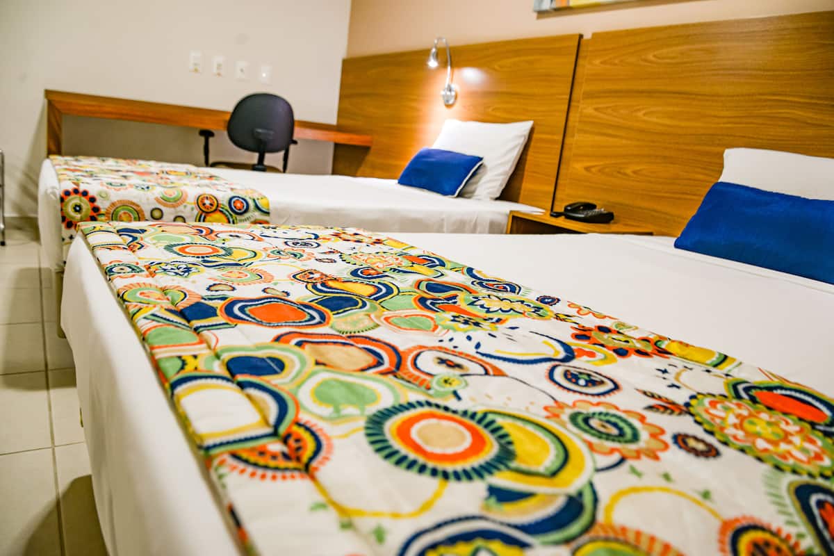 Ein Hotelzimmer mit zwei Betten, jeweils mit bunter, gemusterter Bettwäsche, einem Holzkopfende, einem Schreibtisch mit Stuhl und einem Nachttisch mit Lampe.