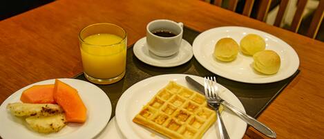 Un desayuno con un gofre, fruta, jugo y café sobre una mesa de madera.