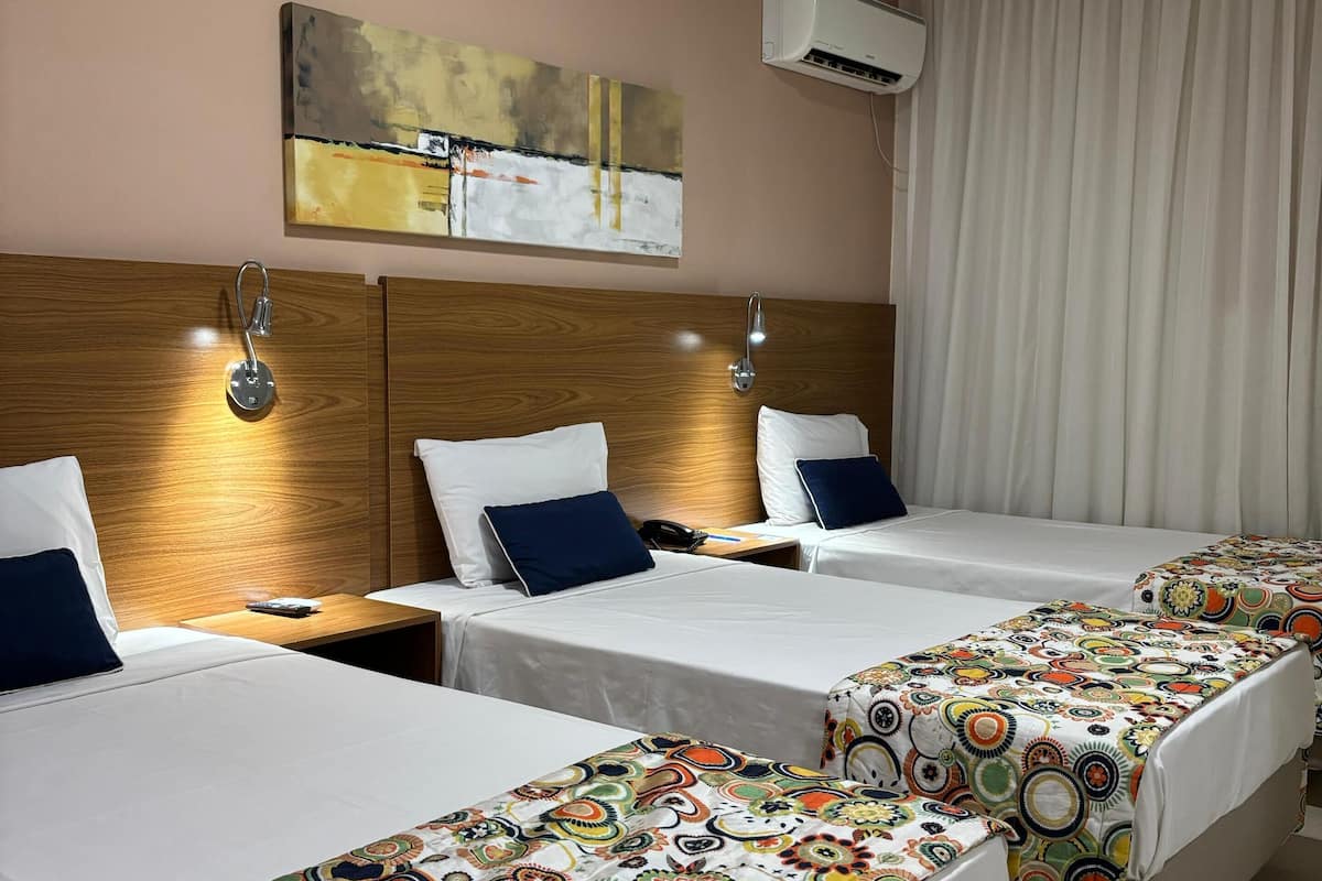 Ein Hotelzimmer mit drei Einzelbetten, jeweils mit farbenfroher, gemusterter Bettdecke. Über den Betten hängt ein Bild an der Wand, eine Klimaanlage ist an der Decke montiert und es gibt ein Fenster mit Vorhängen.