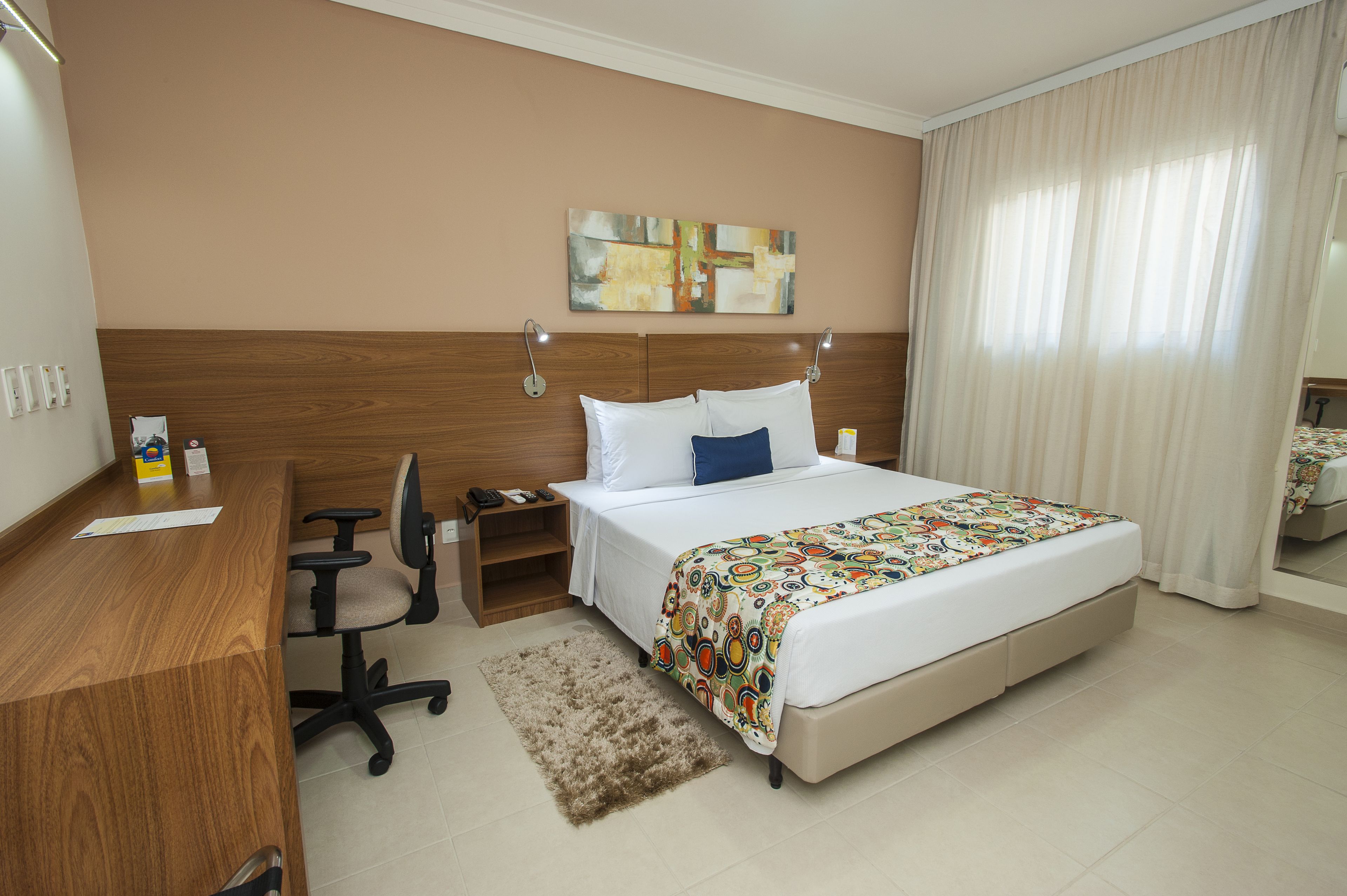 A hotel room with a wooden desk, a bed with a colorful blanket, a nightstand, and a wall-mounted artwork.