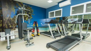 Une salle de sport équipée de tapis de course, d’appareils de musculation et ornée d’une fresque murale représentant une scène de plage.