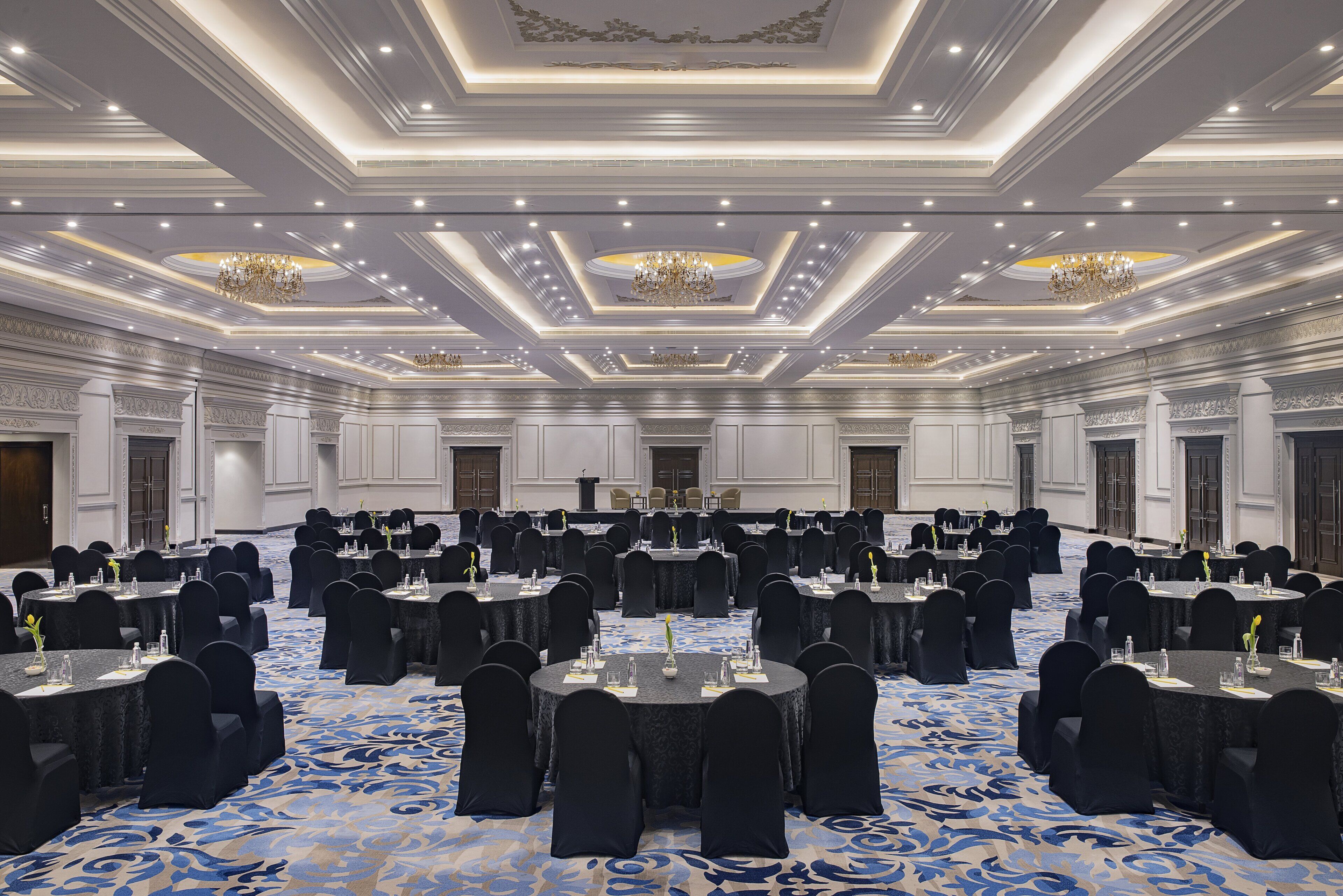 A large banquet hall with round tables, black chairs, and chandeliers.