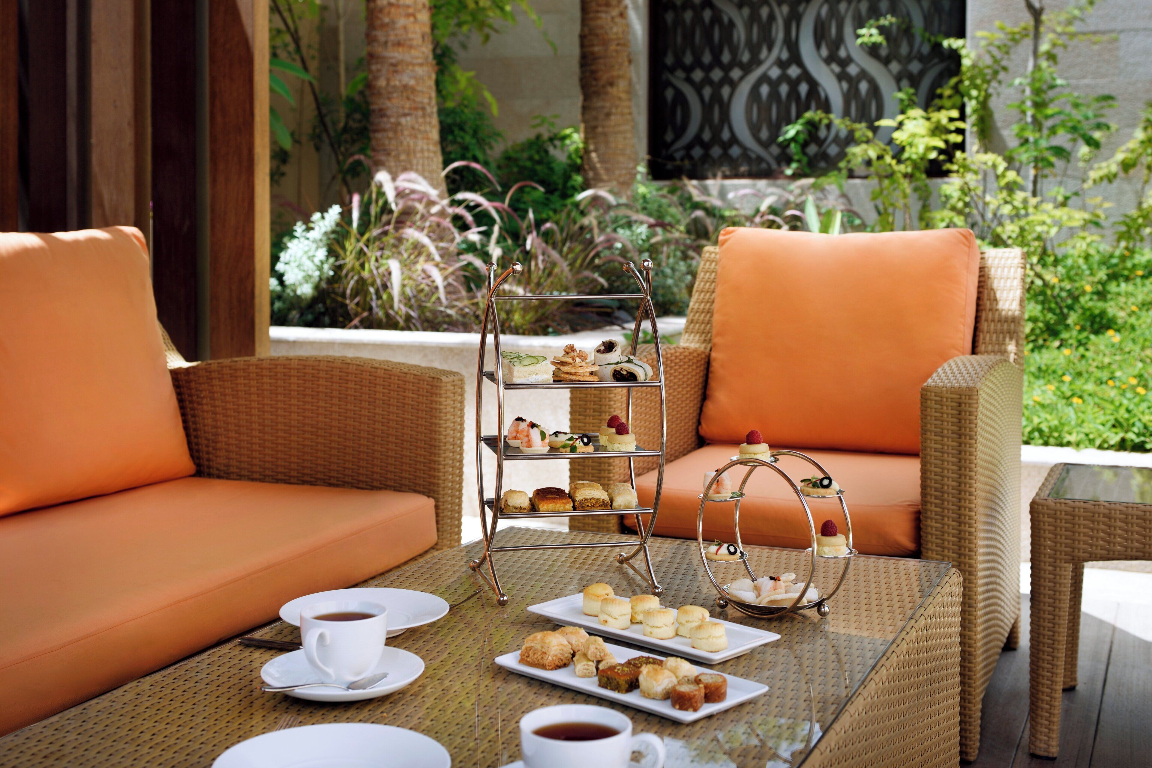 Un sofá de mimbre con cojines naranja, un frutero de tres niveles con pasteles, y una taza de café sobre una mesa.