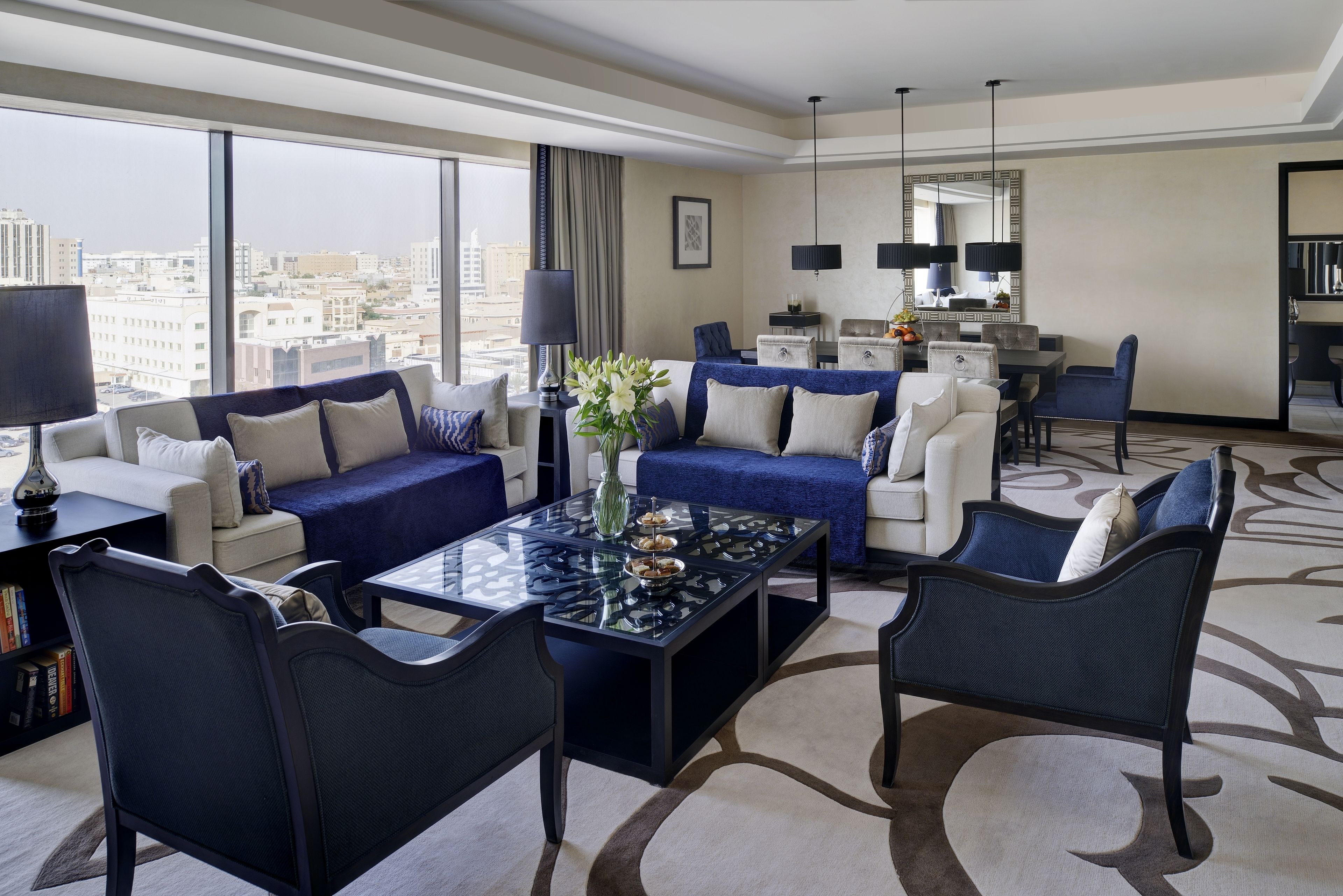 A modern hotel room with a large window offering a city view, a dining area with a table and chairs, and a seating area with sofas and armchairs.