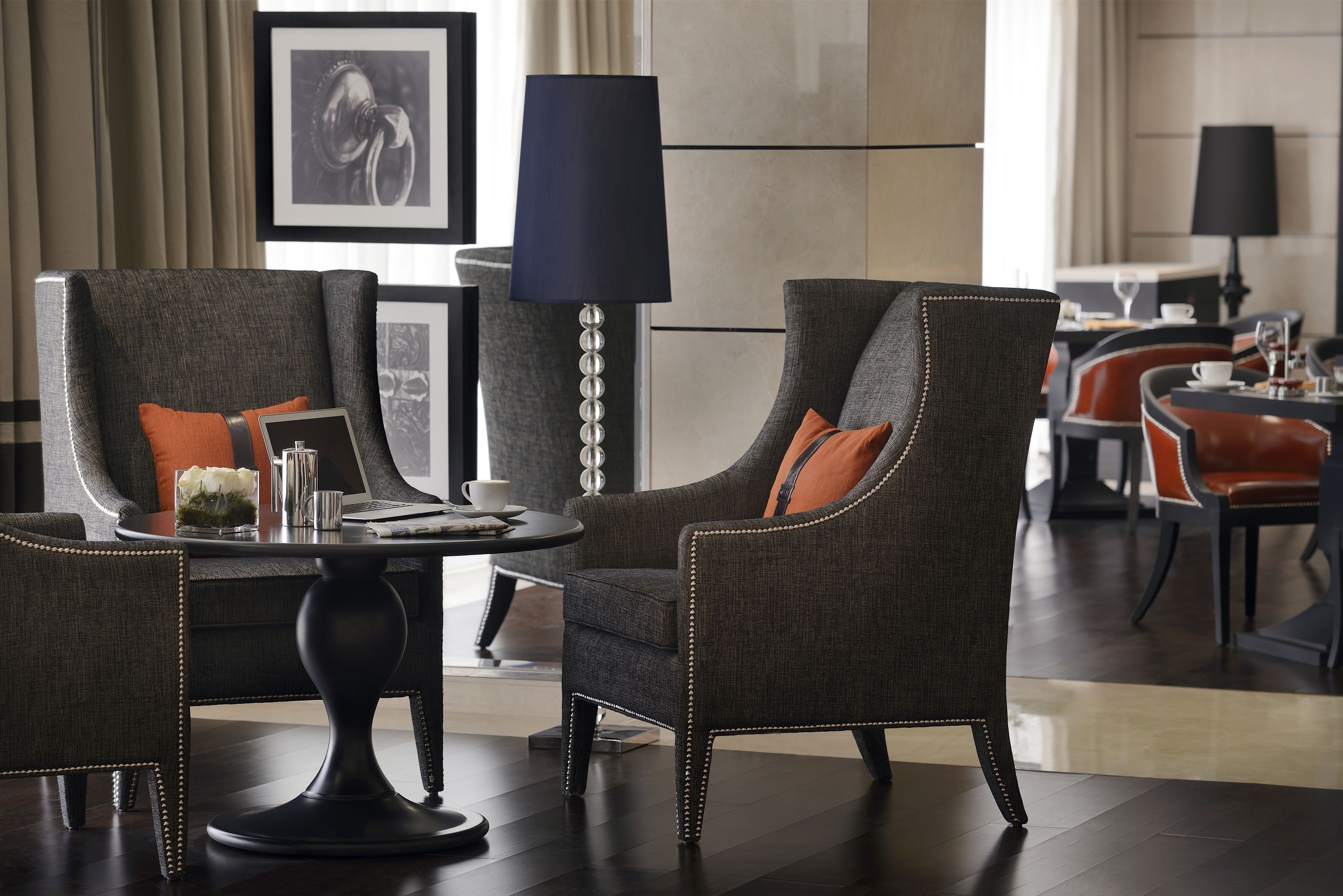 A modern hotel lobby with dark wood flooring, grey upholstered armchairs with orange cushions, a round black table, and a tall floor lamp.