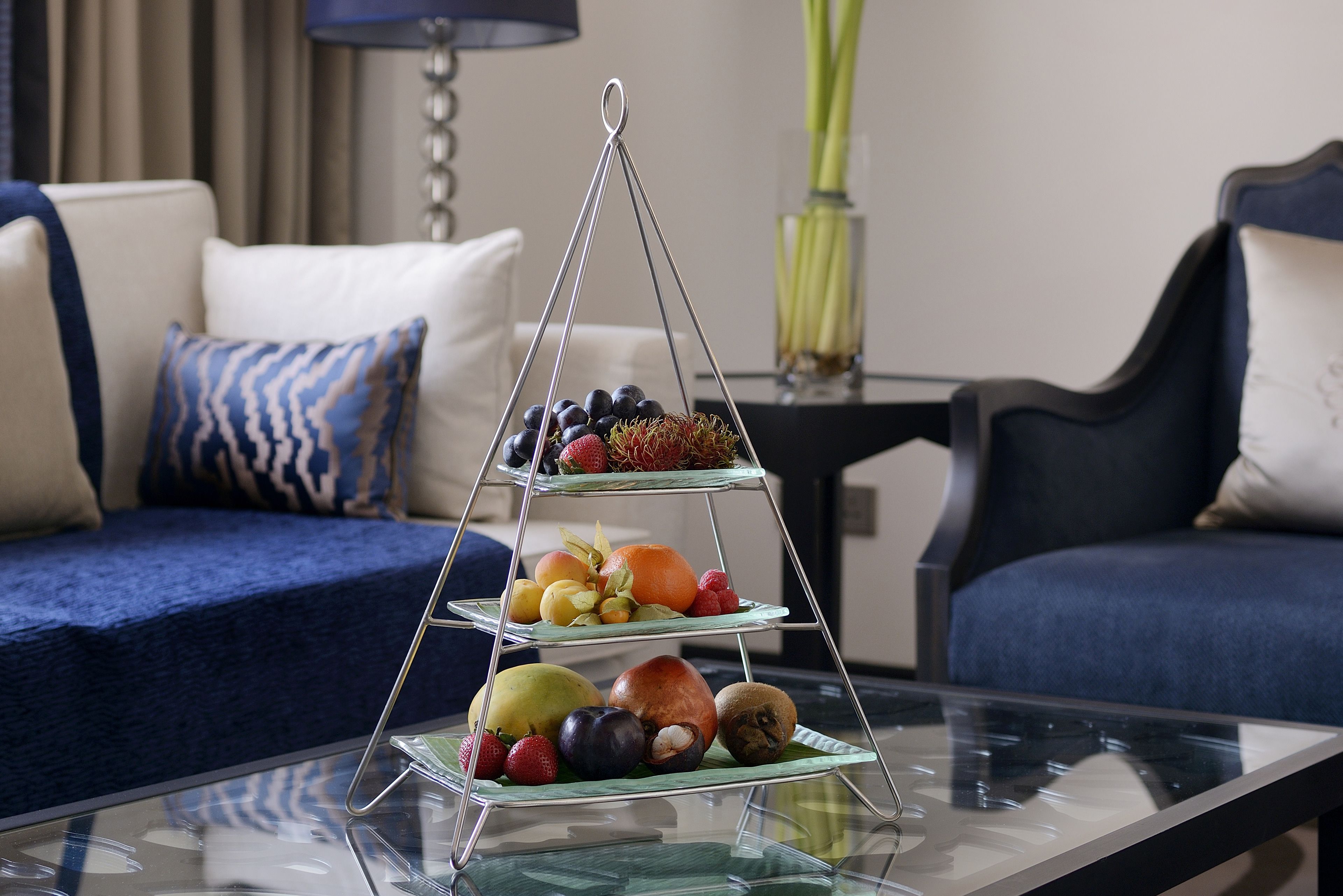 A three-tiered fruit stand on a glass table in a living room with a blue sofa and a vase of flowers in the background.