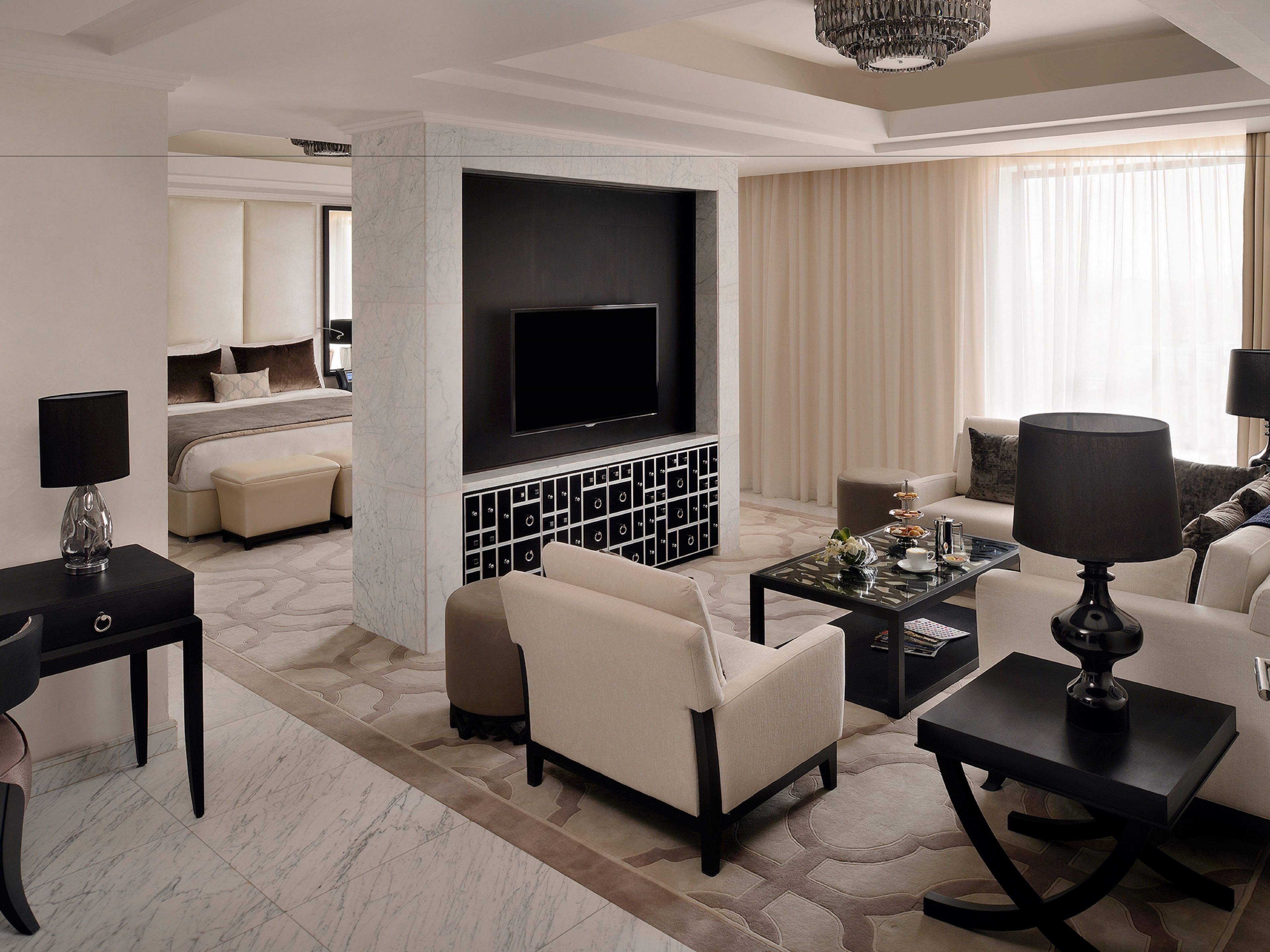 A modern hotel room with a large flat-screen TV, a comfortable sofa, a coffee table, and a dining area with a chandelier.