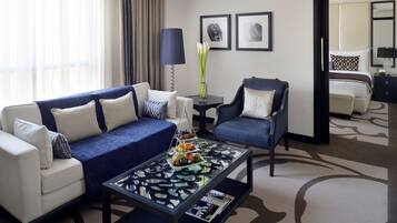 A modern hotel room with a blue sofa, a chair, a coffee table with fruit, and a bed in the background.