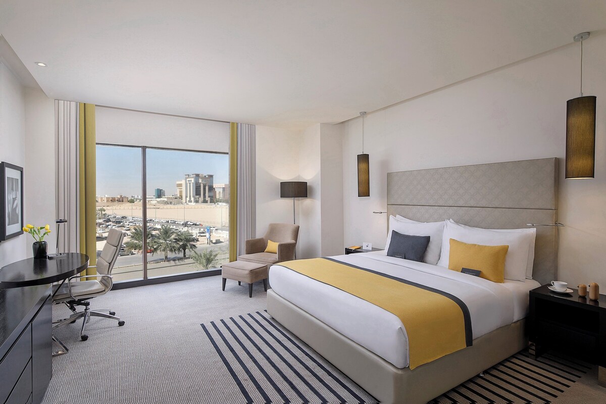 A hotel room with a large bed, a desk, a chair, a lamp, and a view of a cityscape.