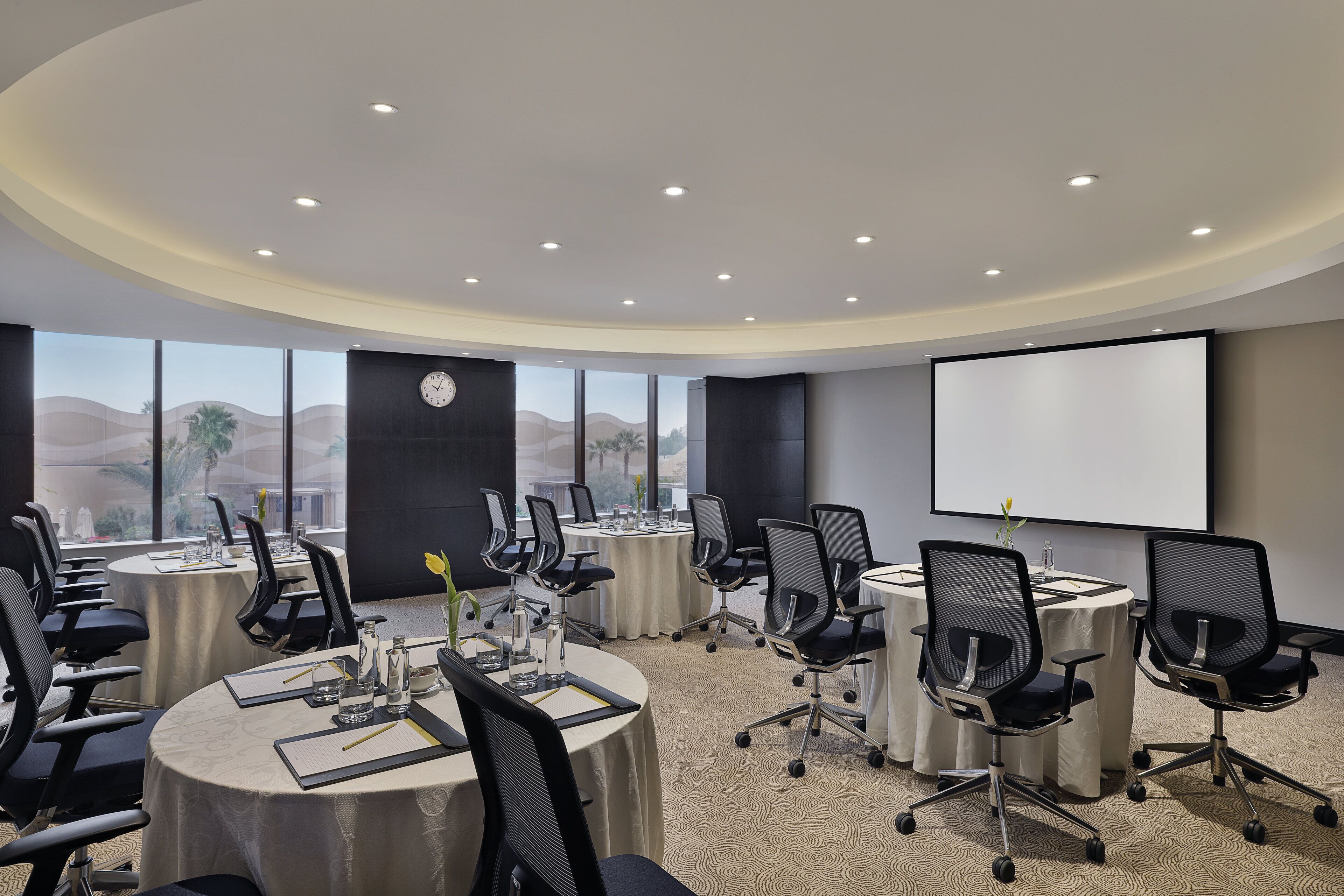 A conference room with round tables, black office chairs, a projector screen, and a clock on the wall.