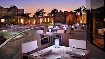 Un espacio de comedor al aire libre con mesas y sillas, una piscina y palmeras.