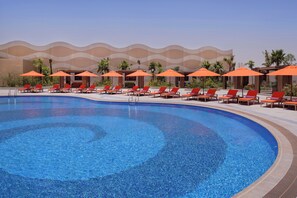 A swimming pool with lounge chairs and umbrellas, surrounded by palm trees and a modern building.