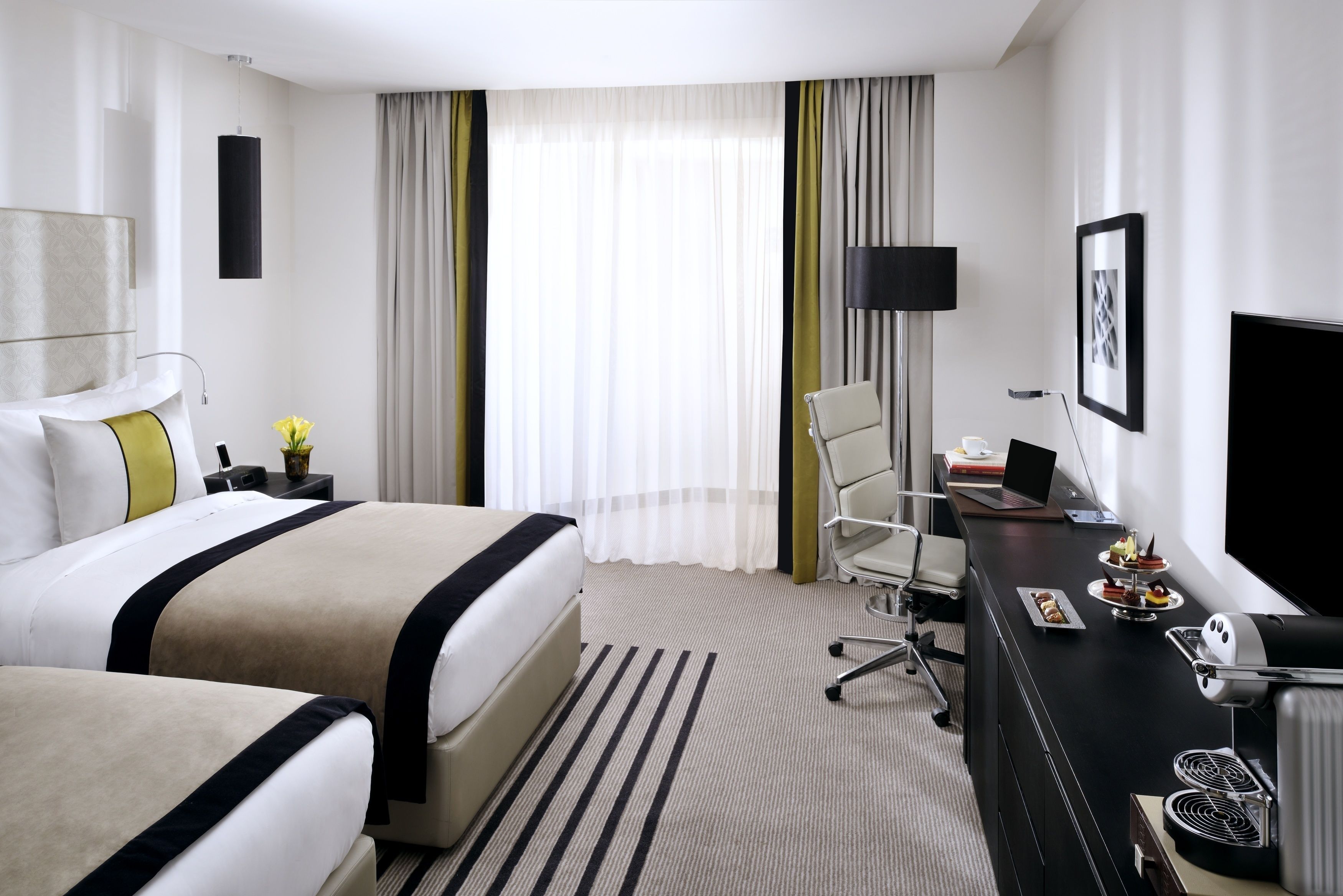 Ett hotellrum med två sängar, ett skrivbord med en bärbar dator, en tv och en matplats med mat.