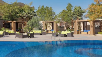 Un espace piscine doté de chaises longues et de parasols, entouré d’arbres et d’un bâtiment.