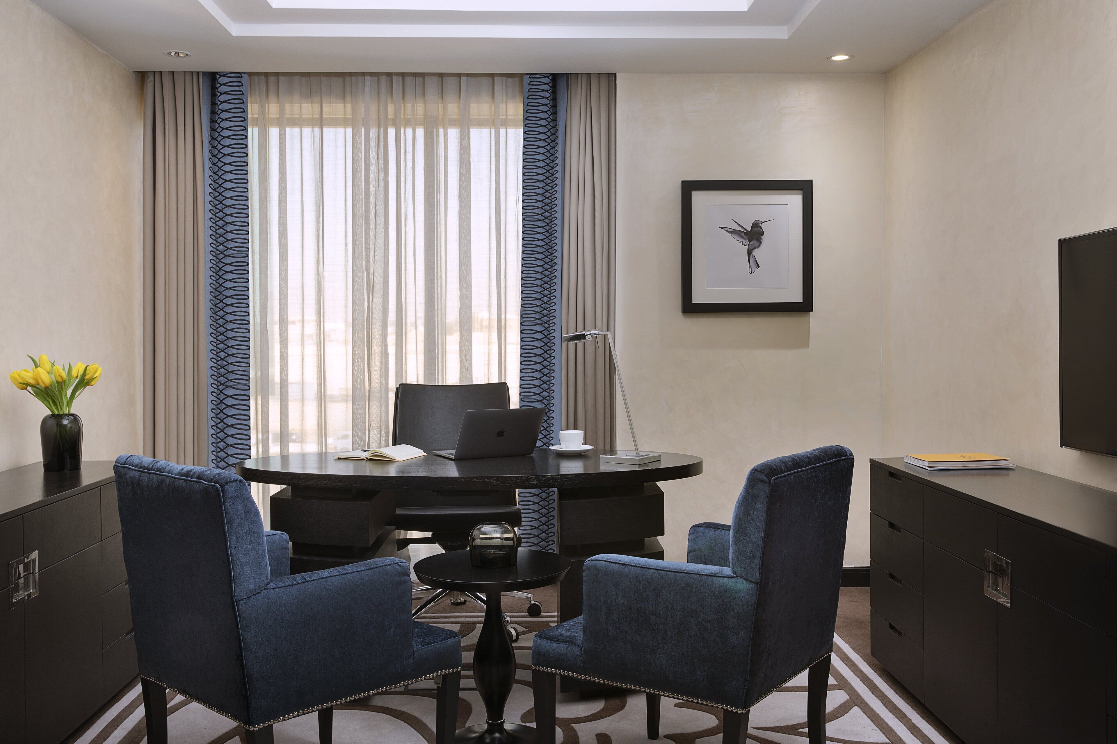 A conference room with a round table, two blue chairs, a laptop, a coffee cup, and a framed picture on the wall.