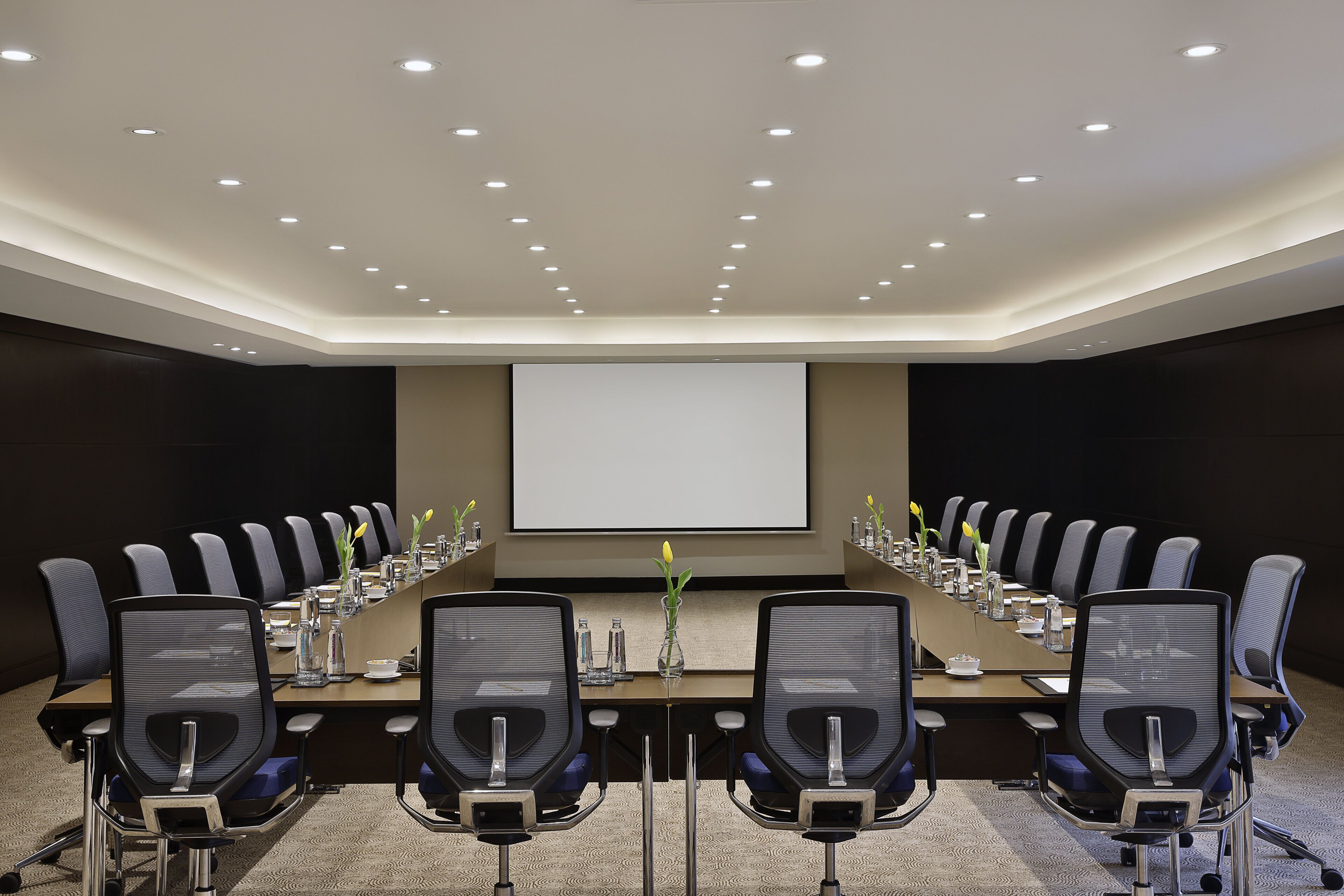 A conference room with a long table, multiple chairs, a projector screen, and a carpeted floor.