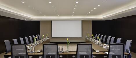 A conference room with a long table, multiple chairs, a projector screen, and a carpeted floor.