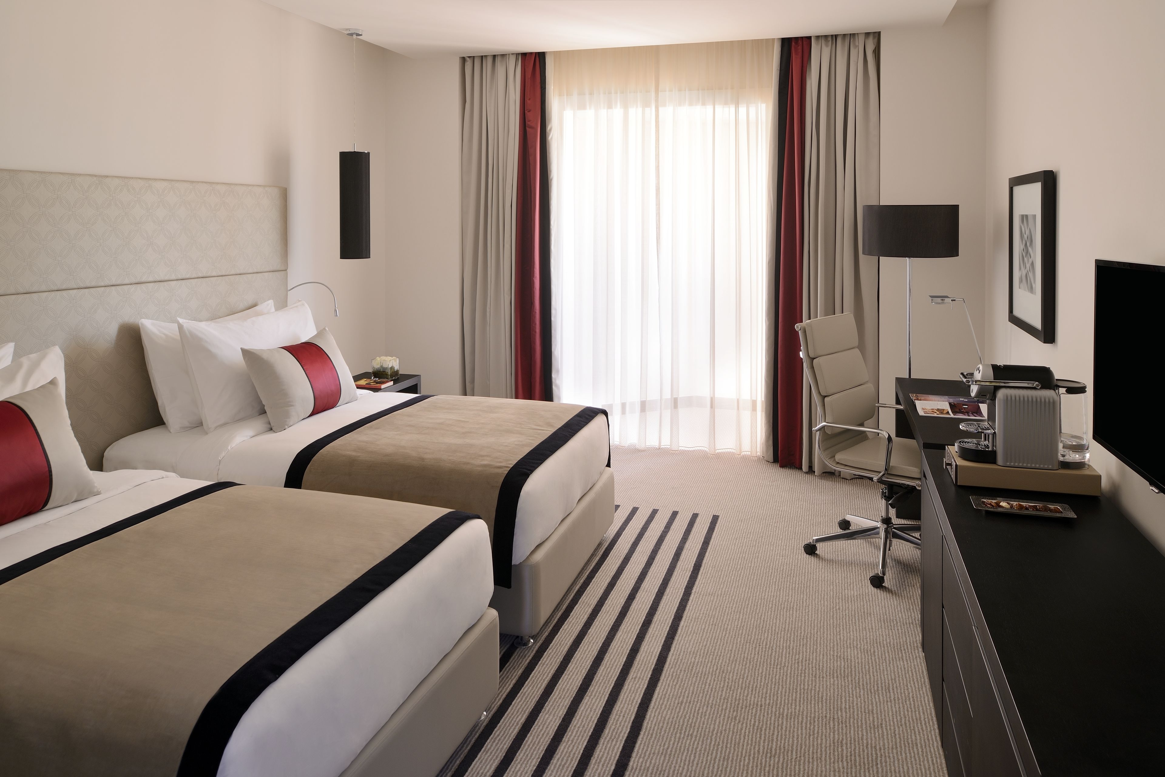 A hotel room with two beds, a desk with a chair, a TV, and a large window with curtains.