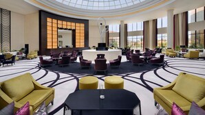 Un hall d’entrée spacieux avec un plafond haut et un espace salon central, comprenant plusieurs canapés et fauteuils, un comptoir d’accueil et de grandes fenêtres laissant entrer la lumière naturelle.