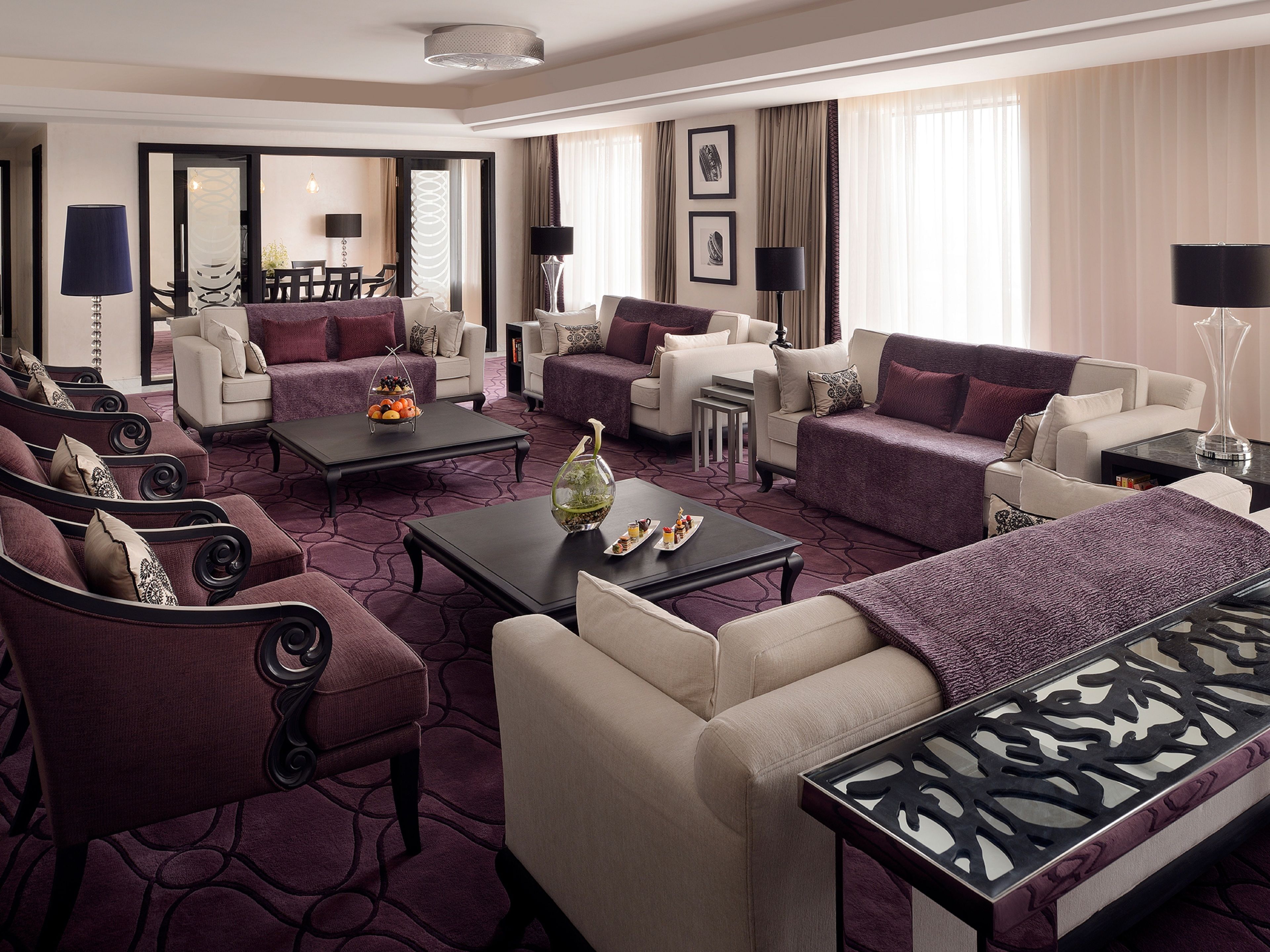 Habitación de hotel moderna con sala de estar que incluye sofás, una mesa de centro y una mesa de comedor. Hay dos lámparas, un jarrón con flores y cuadros en las paredes.